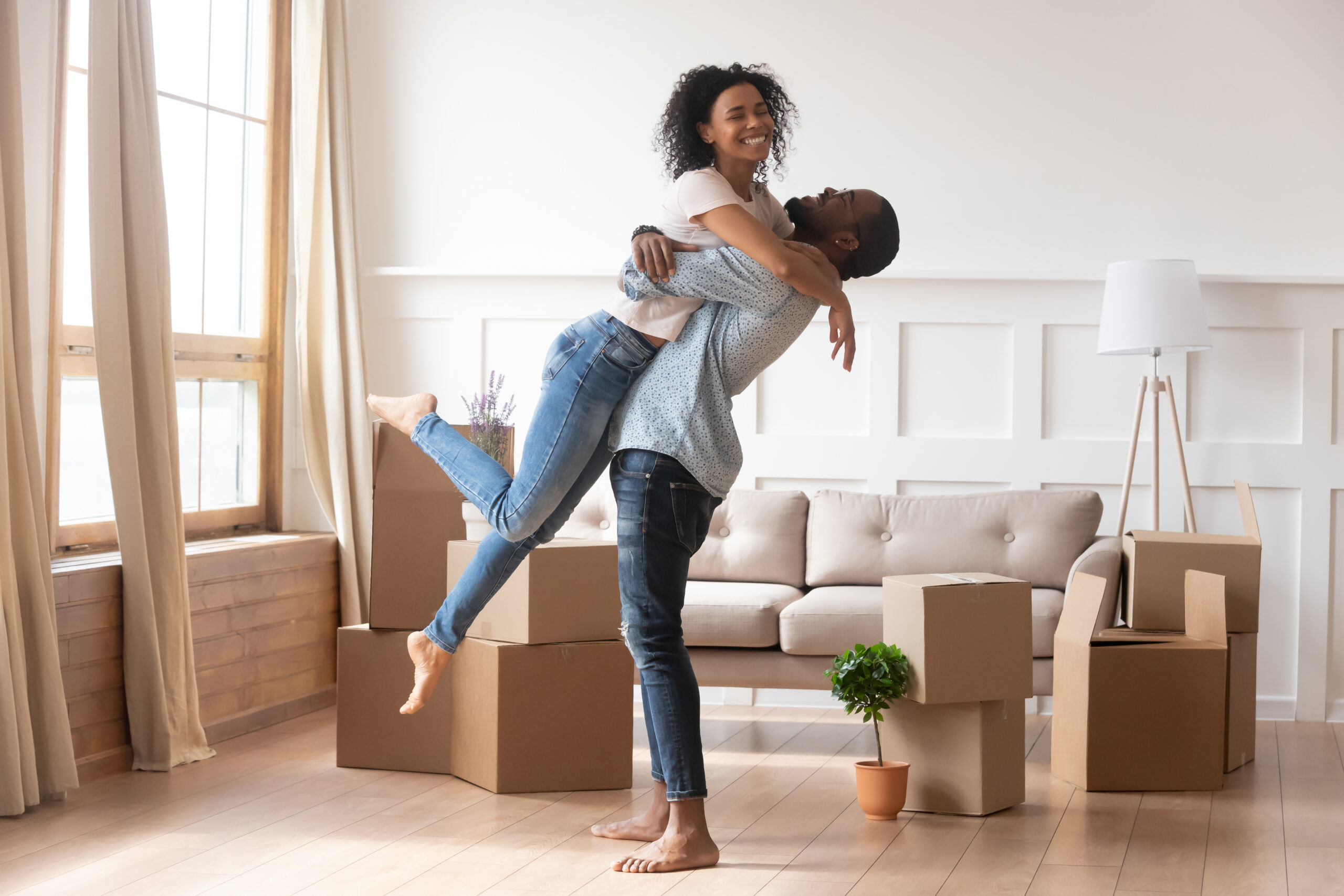 Happy african american couple first time home buyers celebrate new house purchase on moving day, smiling husband embracing lifting excited young wife laughing standing among boxes in own flat house
